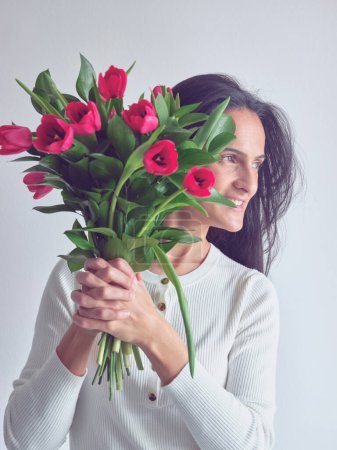 Téléchargez les photos : Portrait de femme heureuse aux cheveux foncés en vêtements décontractés debout avec bouquet de tulipes rouges dans les mains et regardant loin sur fond blanc - en image libre de droit