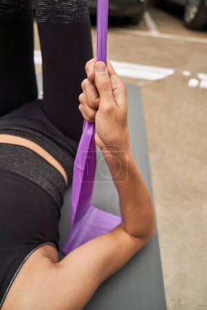 Téléchargez les photos : Athlète féminine méconnaissable en vêtements de sport couché sur tapis de sport gris et les muscles de pompage en utilisant la bande de résistance violette pendant l'entraînement dans le centre de remise en forme moderne - en image libre de droit