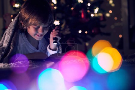 Foto de Libro de lectura de niño encantado con linterna mientras está acostado debajo de la manta en habitación oscura con bokeh colorido y luces brillantes brillantes - Imagen libre de derechos