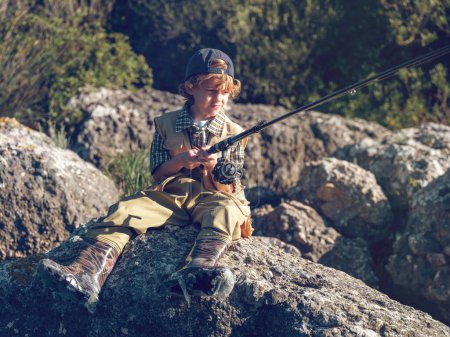 Photo for Full body serious boy in casual clothes and cap looking away and fishing while sitting on stony shore on sunny day in countryside - Royalty Free Image