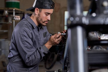 Photo for Side view of concentrated Hispanic male mechanic in uniform carefully fixing details on motorbike wheel in garage with various professional tools - Royalty Free Image