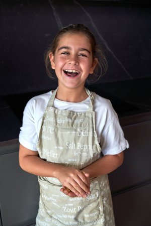 Foto de Niño hispano positivo en delantal mirando a la cámara mientras está de pie en la cocina con poca luz - Imagen libre de derechos