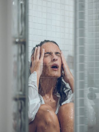 Téléchargez les photos : Femme bouleversée avec les cheveux foncés en chemise mouillée blanche pleurant alors qu'elle était assise dans la douche sous un ruisseau d'eau avec les yeux fermés et la tête touchante - en image libre de droit