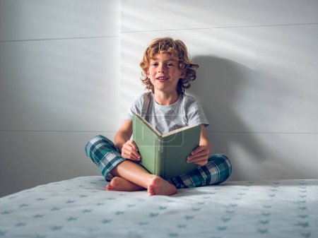 Téléchargez les photos : Préadolescent souriant en vêtements de nuit assis sur un lit confortable et un livre de lecture en journée - en image libre de droit