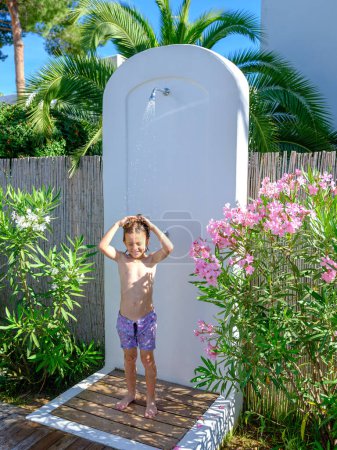 Téléchargez les photos : Corps complet de joyeux petit garçon avec torse nu dans une courte douche dans un jardin tropical par une journée ensoleillée pendant les vacances d'été - en image libre de droit