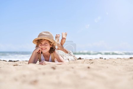 Foto de Cuerpo completo de niño preadolescente alegre en sombrero de paja apoyado en la mano mientras está acostado en la playa de arena cerca del mar y mirando a la cámara durante las vacaciones de verano - Imagen libre de derechos