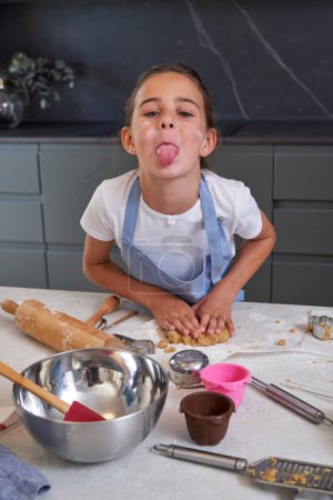 Photo for Hispanic child preparing dough for cookies on table in kitchen and looking at camera with tongue out - Royalty Free Image