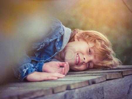 Foto de Niño feliz en chaqueta de mezclilla con el pelo rubio rizado sonriendo y mirando a la cámara mientras está acostado en camino de madera en la naturaleza - Imagen libre de derechos