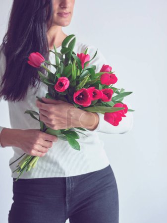 Téléchargez les photos : Recadrer dame anonyme toucher et renifler tas de tulipes lumineuses avec sourire agréable sur le visage tout en se tenant près de fond blanc - en image libre de droit