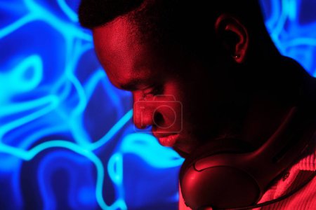 Foto de Vista lateral de cultivo músico masculino afroamericano con auriculares inalámbricos contra la iluminación de neón colorido - Imagen libre de derechos