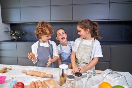 Téléchargez les photos : Enfants gais dans des tabliers pétrissant la pâte pour les biscuits à la table salissante avec divers ingrédients dans la cuisine moderne légère à la maison - en image libre de droit