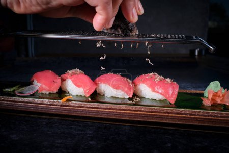 Photo for Crop anonymous cook grating truffle on delicious sushi nigiri made from rice and slices of salmon decorated with radish placed on wooden tray on black background - Royalty Free Image