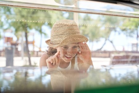 Photo for Through glass view of cheerful curly haired boy in straw straw hat standing near transparent showcase and choosing ice cream in park during warm sunny day - Royalty Free Image
