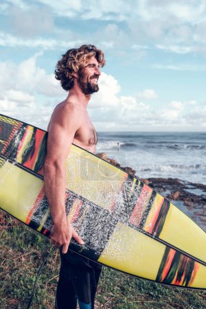 Photo for Side view of cheerful fit Hispanic guy with beard and naked torso smiling brightly while standing on grassy seashore with surfboard and looking away - Royalty Free Image
