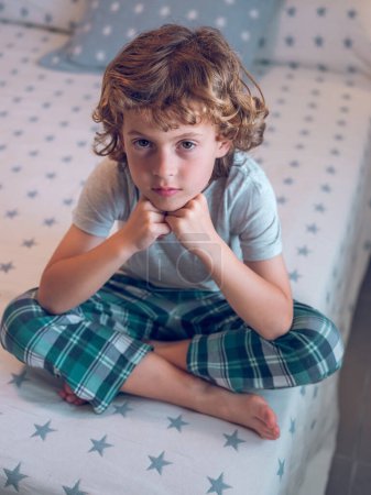 Photo for Full body of adorable curly haired little boy wearing pajamas sitting with crossed legs on bed and looking at camera - Royalty Free Image