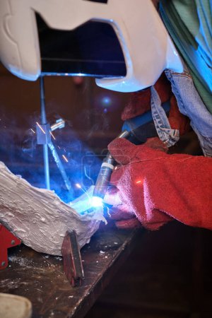 Foto de Vista lateral de un trabajador industrial irreconocible con casco protector y guantes usando linterna de soldadura mientras trabaja en un taller profesional - Imagen libre de derechos