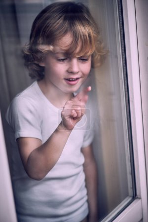 Téléchargez les photos : Grand angle à travers le verre du petit garçon excité avec les cheveux blonds bouclés en t-shirt blanc souriant tout en dessinant sur la condensation de fenêtre à la maison - en image libre de droit