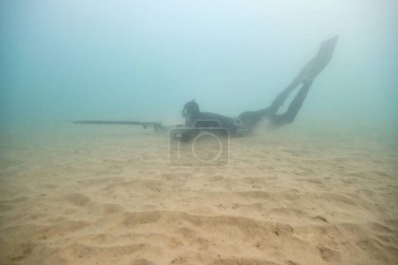 Téléchargez les photos : Vue latérale complète du corps de l'homme anonyme sous-marin en combinaison palmes et lunettes de plongée chasse avec harpon contre la mer floue - en image libre de droit