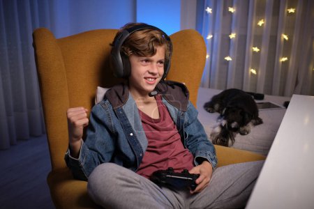 Photo for Positive preteen boy in headset sitting on comfortable armchair with gamepad and celebrating achievement in videogame near bed with dog - Royalty Free Image