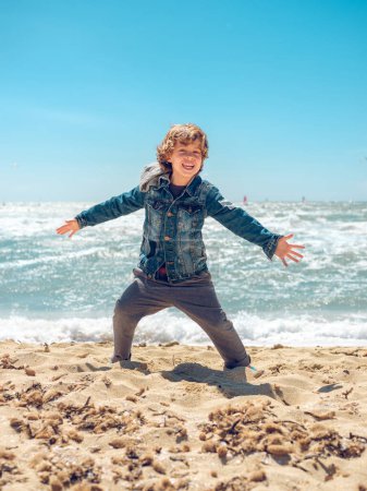 Téléchargez les photos : Corps complet d'enfant heureux avec les cheveux blonds debout sur la plage de sable près de la mer avec les bras tendus et regardant la caméra - en image libre de droit