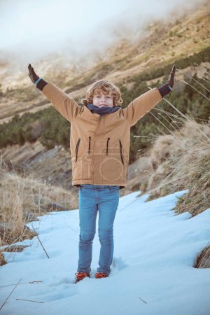 Photo for Cheerful boy in outwear standing with outstretched arms and enjoying winter in snowy highlands while looking at camera - Royalty Free Image