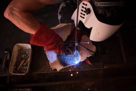 Photo for From above of anonymous craftsman in protective helmet and gloves standing at workbench with torch and welding metal details in gypsum form while working in dark professional workshop - Royalty Free Image