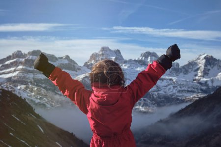 Foto de Vista posterior de un niño irreconocible levantando brazos y celebrando el éxito mientras está parado contra la cordillera nevada y el cielo azul nublado en un día frío en la naturaleza - Imagen libre de derechos