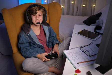 Hoher Winkel des entzückenden Jungen mit Steuerknüppel, der auf einem Stuhl sitzt und in die Kamera schaut, während er zu Hause Videospiele spielt