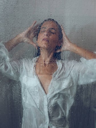 Téléchargez les photos : À travers le verre de femme attrayante en chemisier avec lavage des cheveux noirs humides dans la cabine de douche légère avec porte transparente dans la salle de bain - en image libre de droit