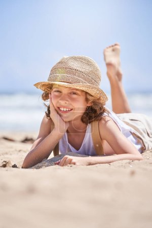 Téléchargez les photos : Rez-de-chaussée d'un enfant souriant en chapeau de paille appuyé sur la main et regardant la caméra alors qu'il était allongé sur une plage de sable près de la mer pendant les vacances d'été - en image libre de droit