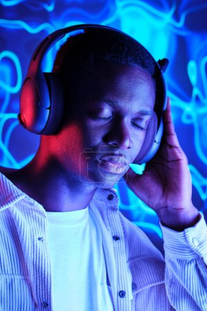 Téléchargez les photos : Homme afro-américain paisible portant des vêtements décontractés écoutant la playlist dans un casque sans fil contre l'éclairage au néon bleu - en image libre de droit