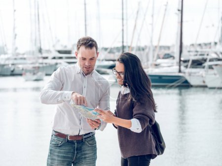 Male pointing on map while searching route for smiling female tourist standing on waterfront during vacation