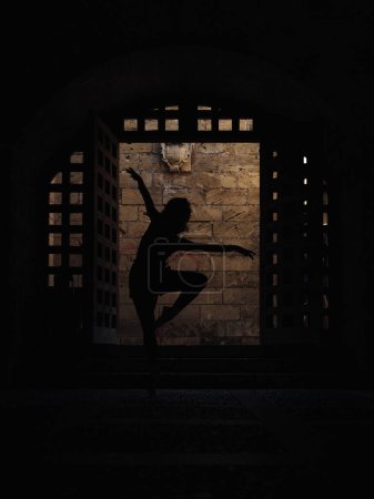 Photo for Silhouette of unrecognizable talented female ballet dancer performing dance movement on dark street near shabby building with doorway in city - Royalty Free Image