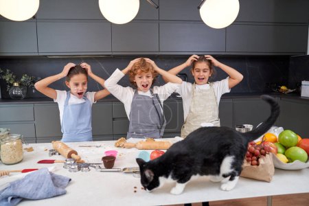 Téléchargez les photos : Enfants étonnés avec les mains sur la tête regardant chat noir sur la table salissante avec des fruits tout en préparant des biscuits dans la cuisine légère - en image libre de droit