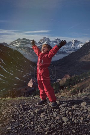 Téléchargez les photos : Enfant en vêtements d'extérieur rouges levant les bras et criant debout contre des montagnes enneigées et ciel bleu nuageux le jour du printemps dans la nature - en image libre de droit