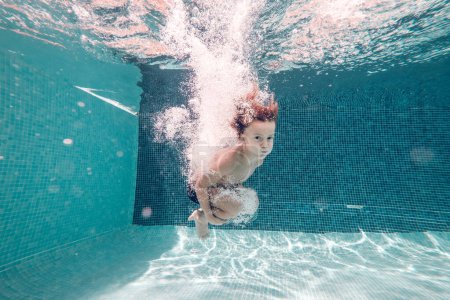 Foto de Vista submarina del niño descalzo con el torso desnudo usando pantalones cortos de natación saltando y buceando en agua transparente clara abrazando las rodillas y mirando a la cámara - Imagen libre de derechos