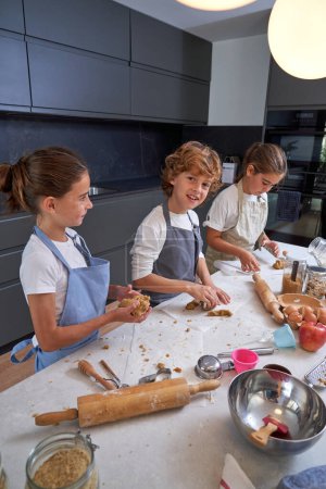 Photo for Group of children in aprons standing at table in modern kitchen and processing dough while preparing cookies - Royalty Free Image