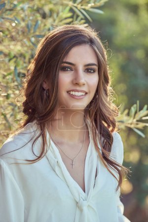 Photo for Charming young female with curly hair in white blouse looking at camera with smile while standing near trees on blurred background - Royalty Free Image