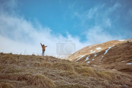 Téléchargez les photos : Garçon lointain méconnaissable en vêtements de dessus debout sur une colline herbeuse avec les bras levés près de la pente avec le givre par temps froid dans la nature - en image libre de droit