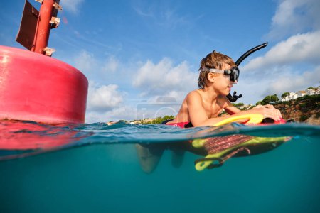 Téléchargez les photos : Vue latérale du préadolescent souriant en masque de plongée et tuba nageant en eau de mer sur une planche flottante près de la bouée de sauvetage rouge près de la plage - en image libre de droit
