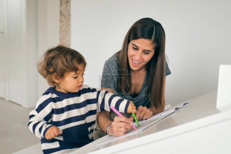 Téléchargez les photos : Femme souriante et curieux petit garçon regardant vers le bas et dessinant avec des stylos en feutre ensemble dans le salon à la maison - en image libre de droit
