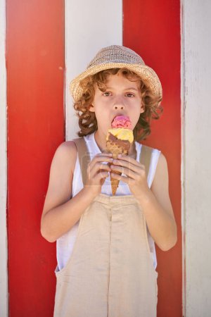 Foto de Adorable niño en sombrero de paja y en general de pie contra el fondo rayado rojo y blanco mientras come helado dulce en el cono de gofre y mirando a la cámara - Imagen libre de derechos