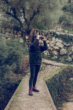 Photo for Full body of young active female traveler with dark hair in casual clothes and sneakers smiling while admiring nature through binoculars standing on narrow path during hiking trip in nature - Royalty Free Image