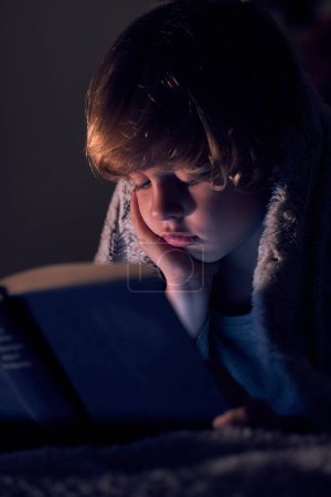 Foto de Chico tranquilo envuelto en una manta caliente mientras lee un libro interesante en la cama en habitación oscura con poca luz en casa - Imagen libre de derechos