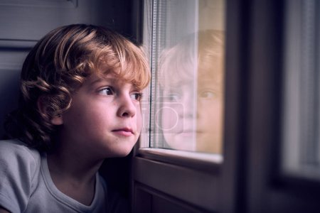 Téléchargez les photos : Adorable petit garçon coûteux aux cheveux blonds bouclés appuyé sur la fenêtre et regardant loin rêveur à la maison - en image libre de droit