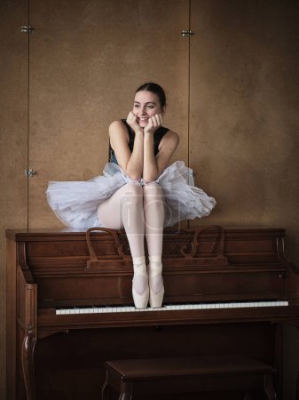 Téléchargez les photos : Corps complet de ballerine féminine en tutu blanc et pointes de chaussures assis sur le dessus du piano et regardant loin heureusement - en image libre de droit