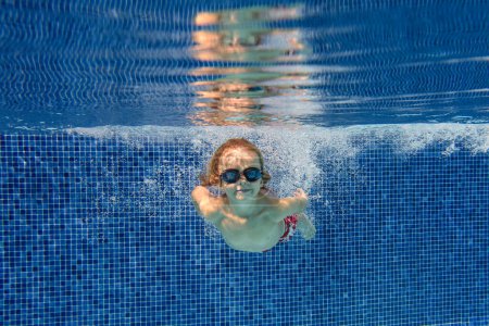 Foto de Vista submarina de un niño adorable con gafas nadando en agua limpia y transparente de la piscina y mirando a la cámara contra la pared azul - Imagen libre de derechos