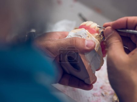 Foto de De arriba de la cosecha técnica dental irreconocible tallado dentadura postiza hecha de dientes de porcelana artificial utilizando un instrumento metálico afilado en el laboratorio - Imagen libre de derechos