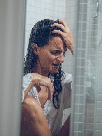 Foto de Vestida dama de pelo largo llorando en cabina de ducha mientras se sienta bajo rociar agua y tocar la frente - Imagen libre de derechos