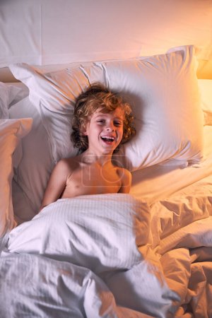 Foto de Vista superior de niño sin camisa emocionado con el pelo rizado acostado debajo de una manta caliente en la cama suave y riendo en el dormitorio tenue en casa - Imagen libre de derechos